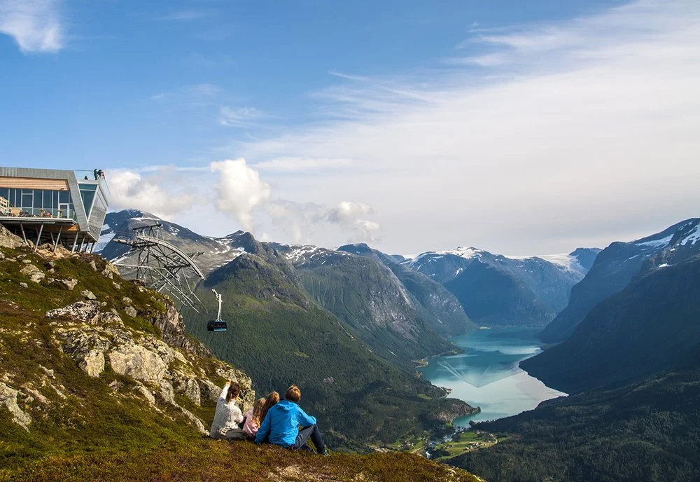 Three people sitting on a grassy slope overlooking a valley with a lake, surrounded by mountains with snow, and a modern building with an observation deck on the left.