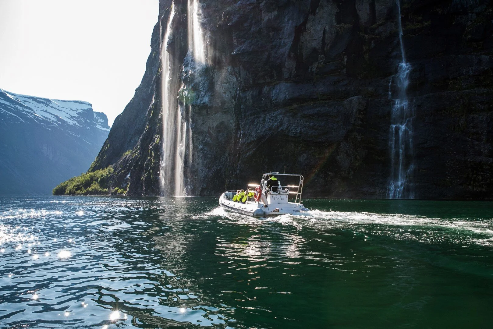 A boat sailing on a fjord with steep cliffs and waterfalls, snow-capped mountains in the background, and sunlight reflecting off the water.