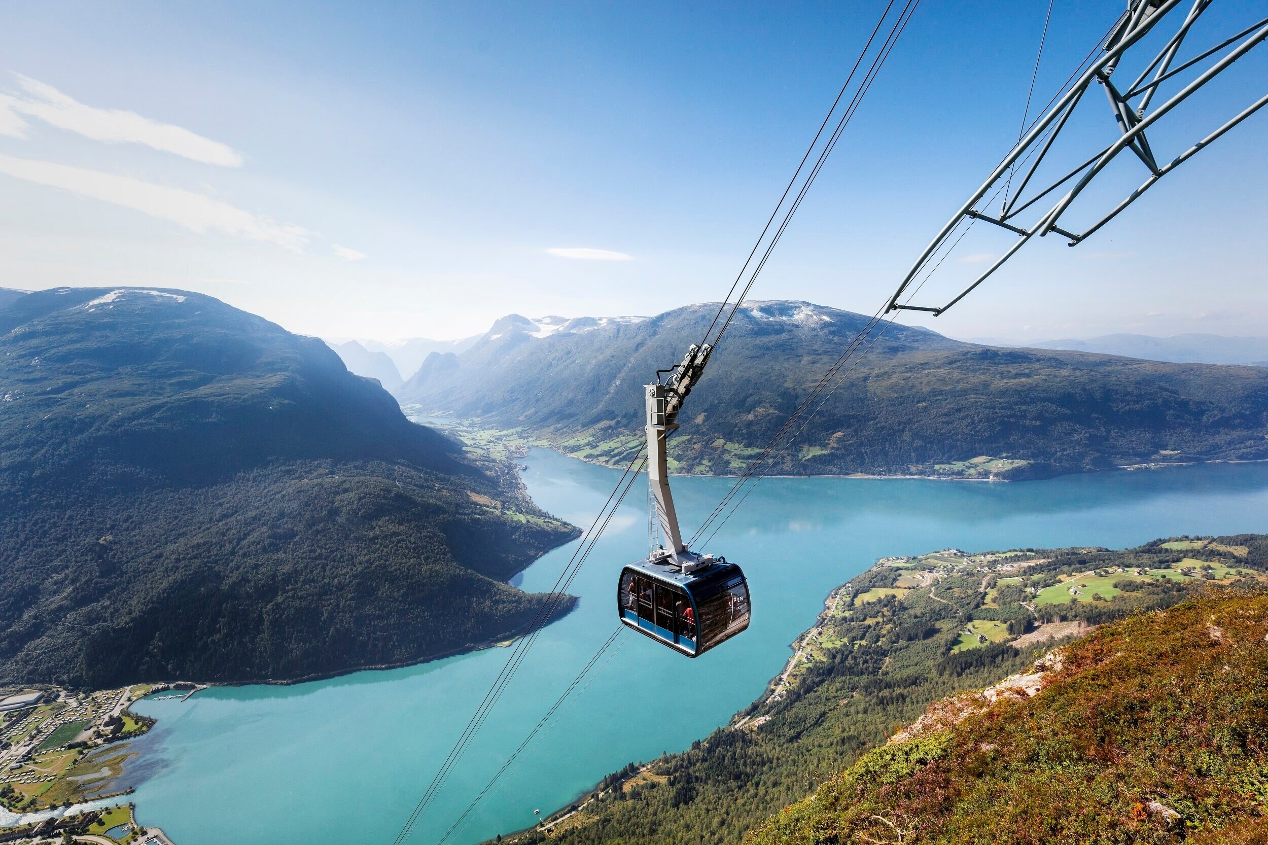 A cable car traveling over a scenic landscape with a large river or lake, surrounded by green mountains and valleys.