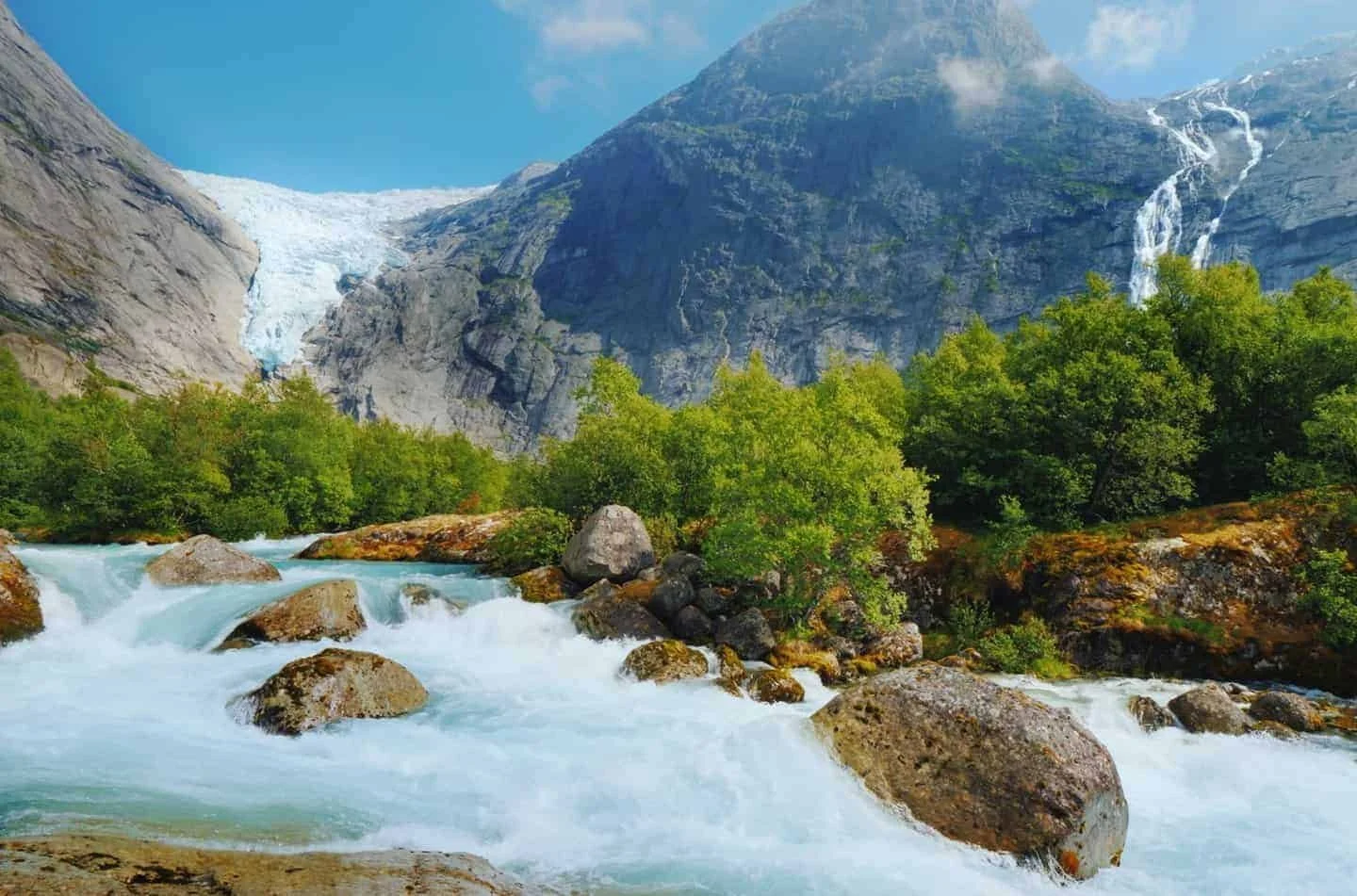 A mountain landscape with a flowing river, lush green trees, and waterfalls on the mountain in the background during daylight.