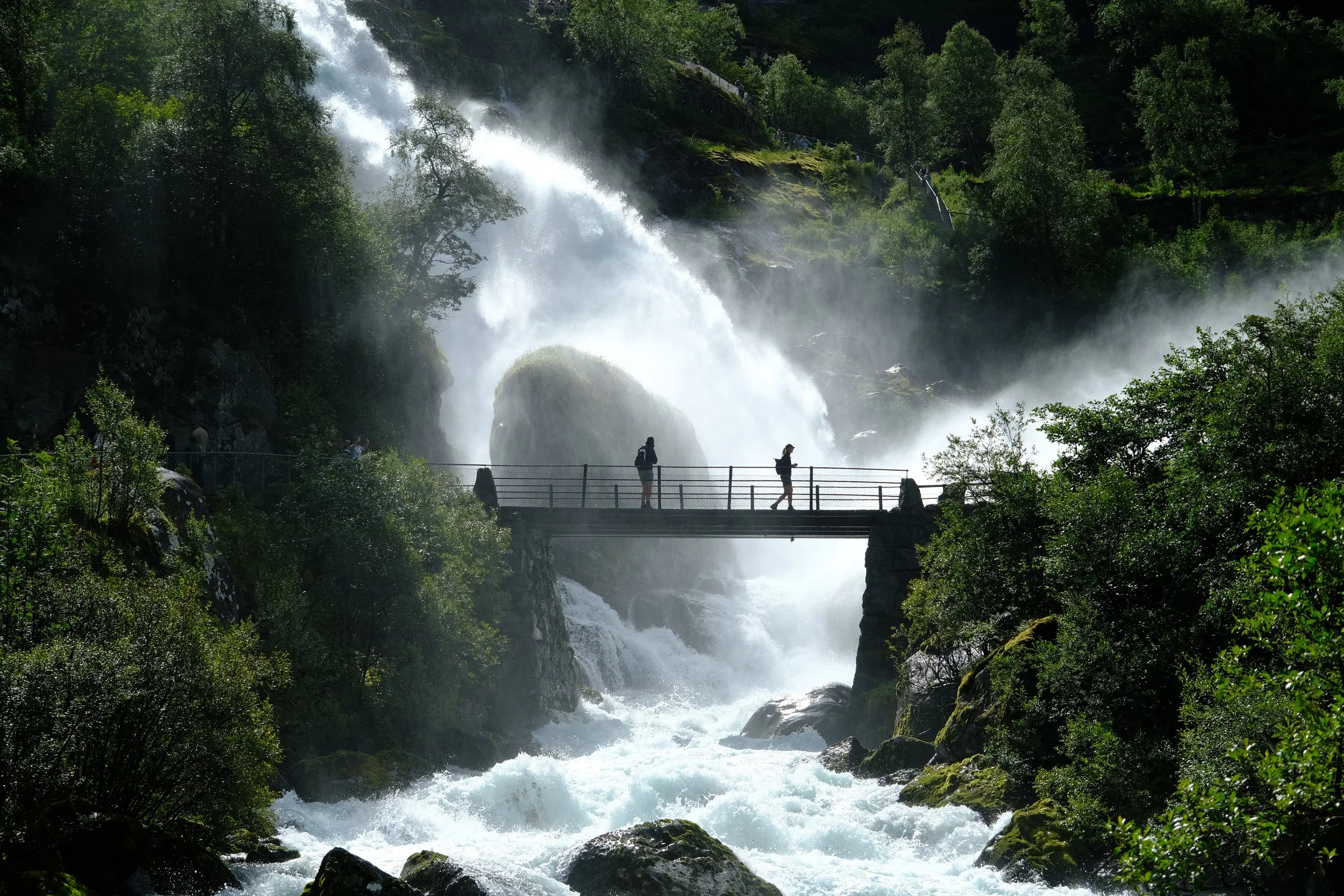 People walking on a bridge over a rushing river in front of a large waterfall amid lush green trees.
