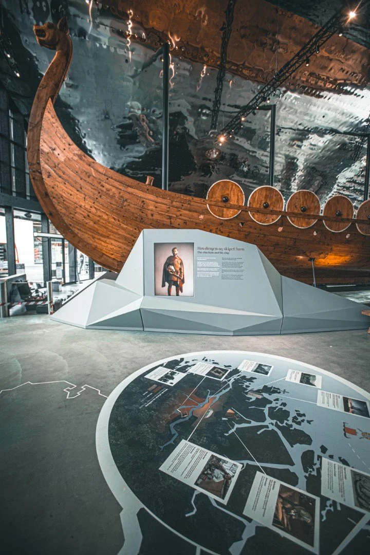 Indoor museum exhibit with a large wooden Viking ship displayed on a platform and informational display panels on the floor and wall.