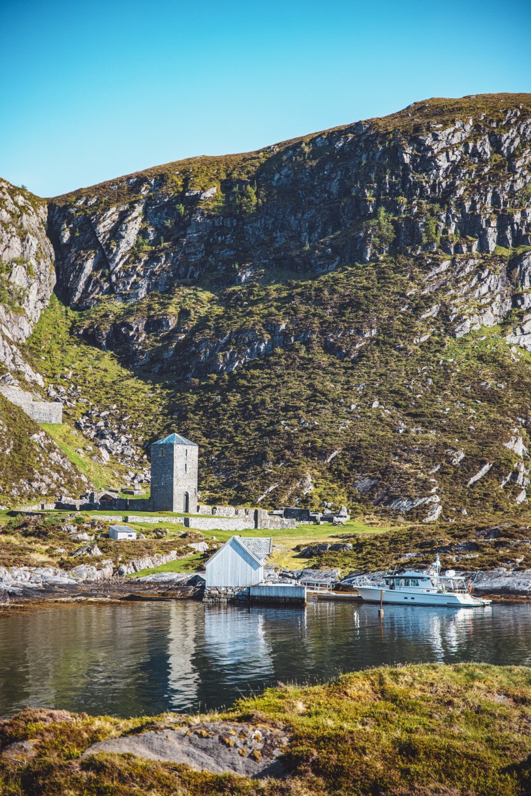 A peaceful harbor scene with a white boat docked near a small wooden boathouse, a stone tower, and a rugged hillside covered in green shrubbery under a clear blue sky.