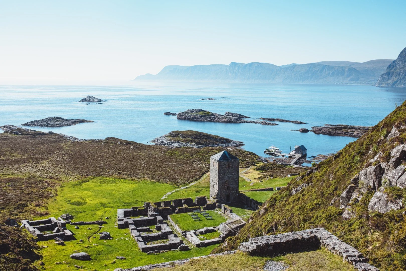 Ruins of an old castle on a green hillside overlooking the coast with rocky islands and mountains in the distance.