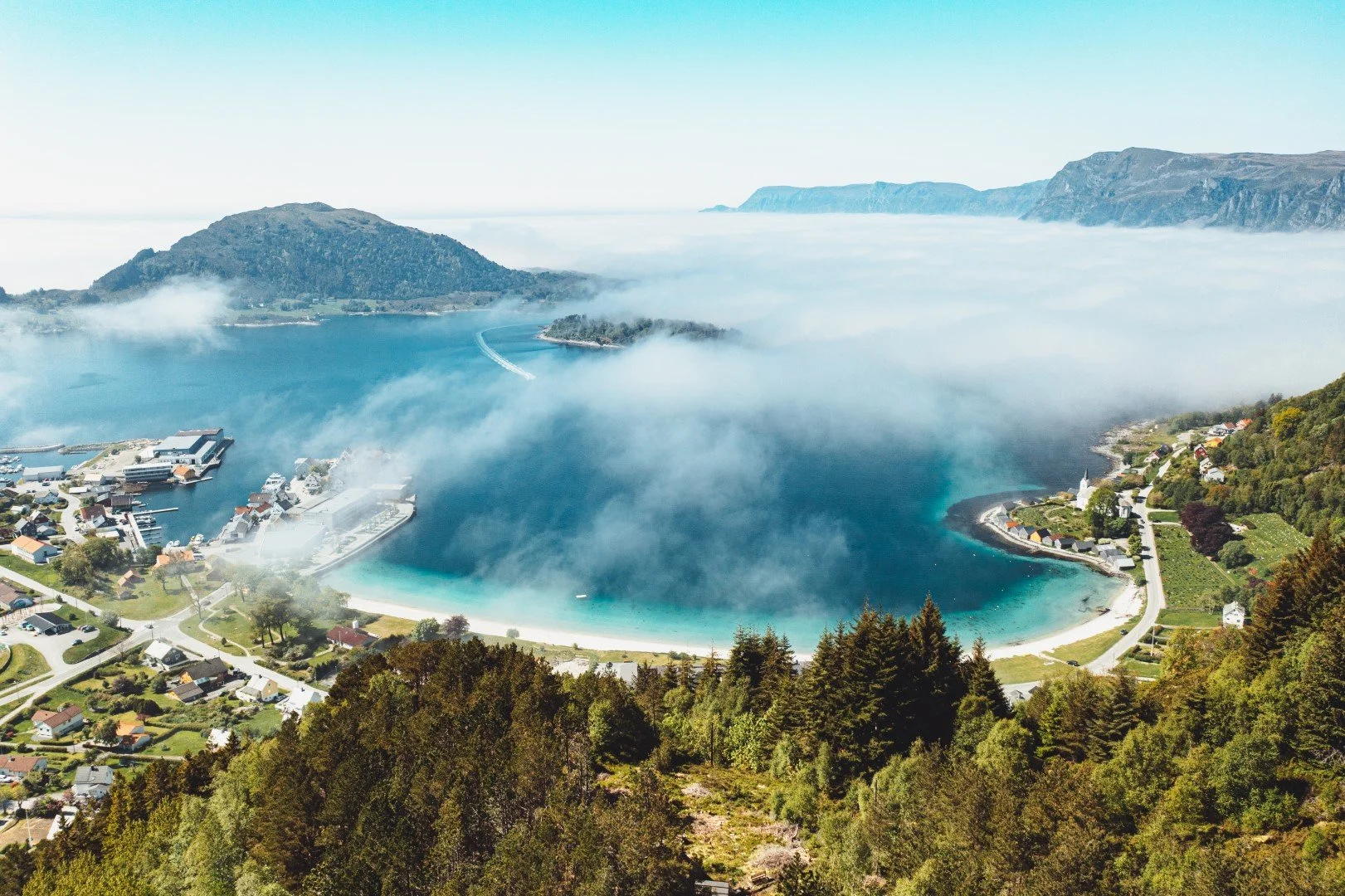 A scenic view of a fjord with a small town along its shoreline, surrounded by green forests and mountains, shrouded in fog with some clear skies above.