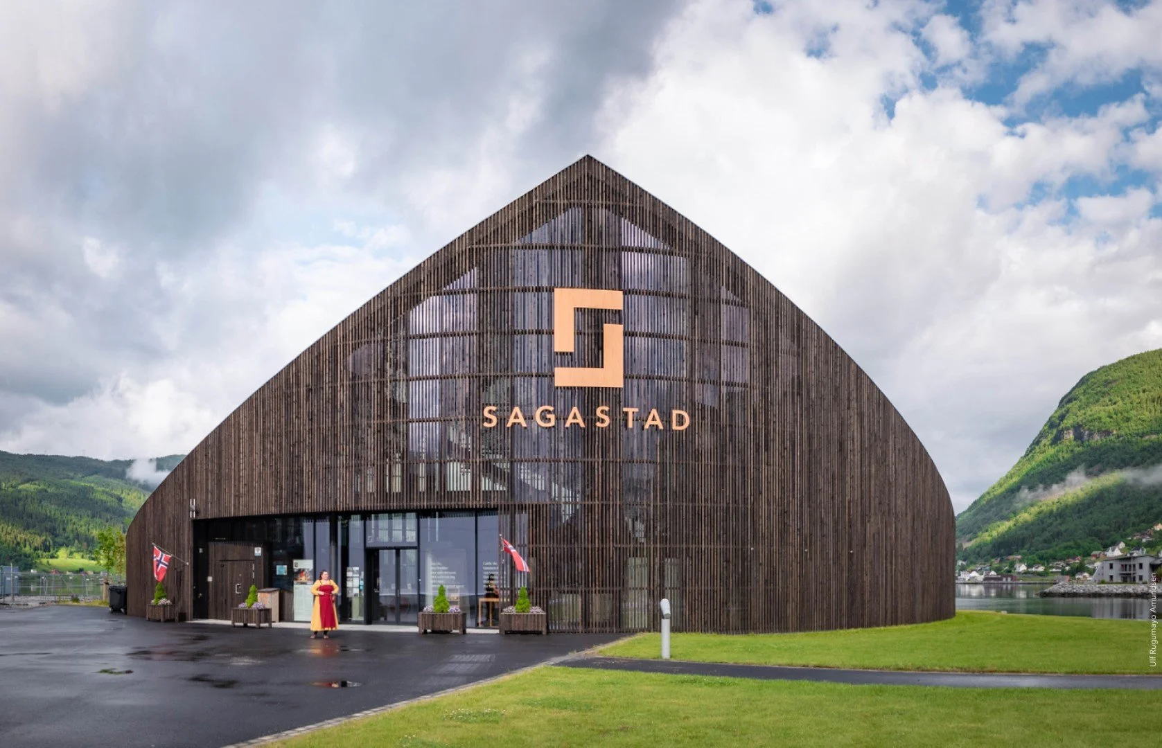 Modern wooden building with the name Sagastad, situated near water and surrounded by green hills and mountains; cloudy sky overhead.