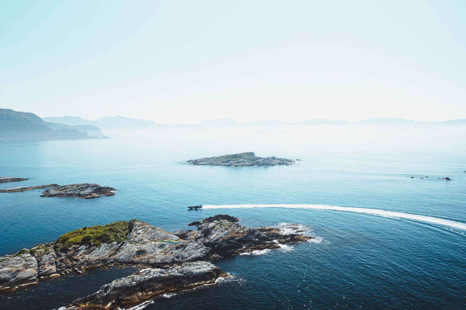 A coastal landscape with rocky islands, calm blue water, and distant mountains, with a boat speeding across the water leaving a wake behind.