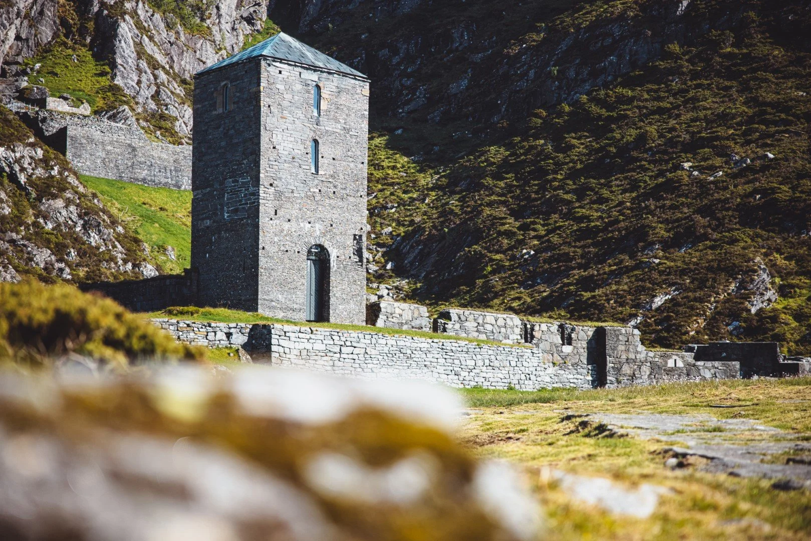 Stone tower with small windows and a door, surrounded by a stone wall, set against a hillside covered with bushes and rocks.