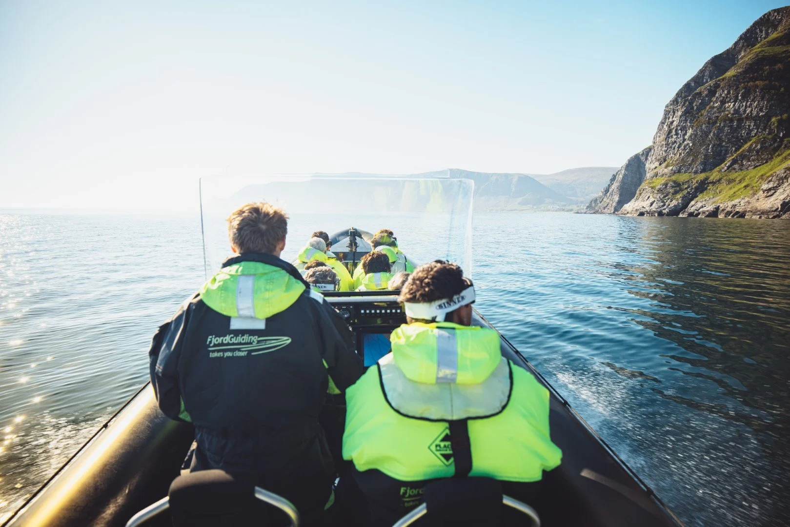 Group of people on a boat touring on a calm lake with cliffs in the background, wearing high-visibility jackets.