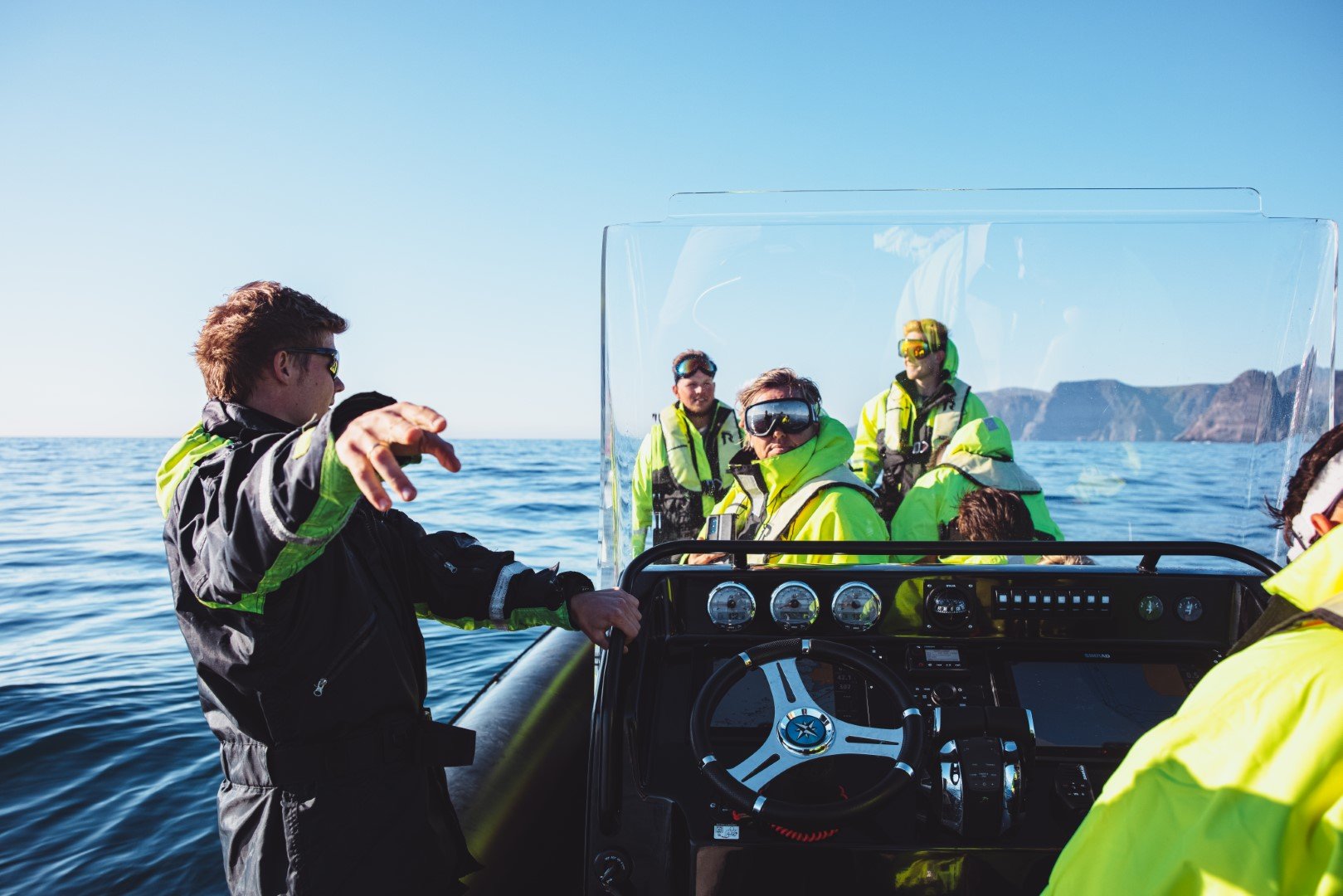 A group of people on a boat wearing yellow and black waterproof jackets, with one person steering and others wearing goggles, during a sunny day on the water with mountains in the background.