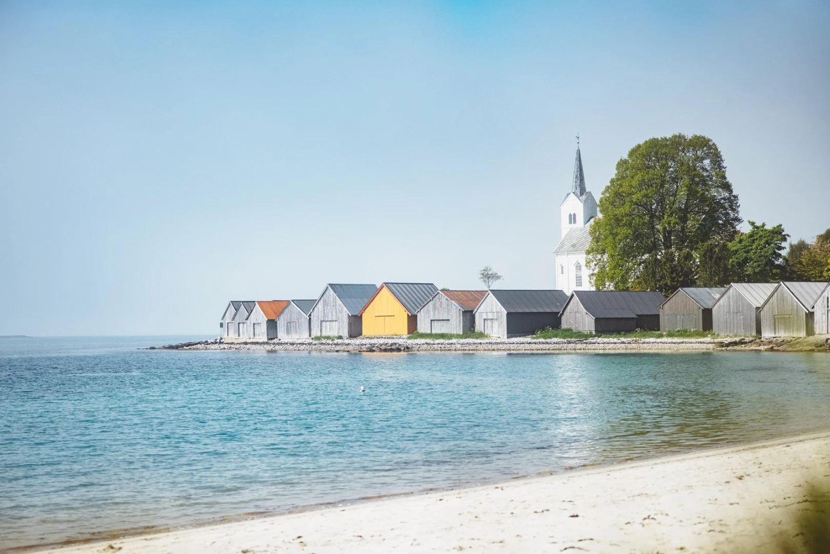 A serene seaside scene with a row of colorful and gray wooden boathouses along the shoreline, a large tree behind the boathouses, and a white church with a steeple in the background.