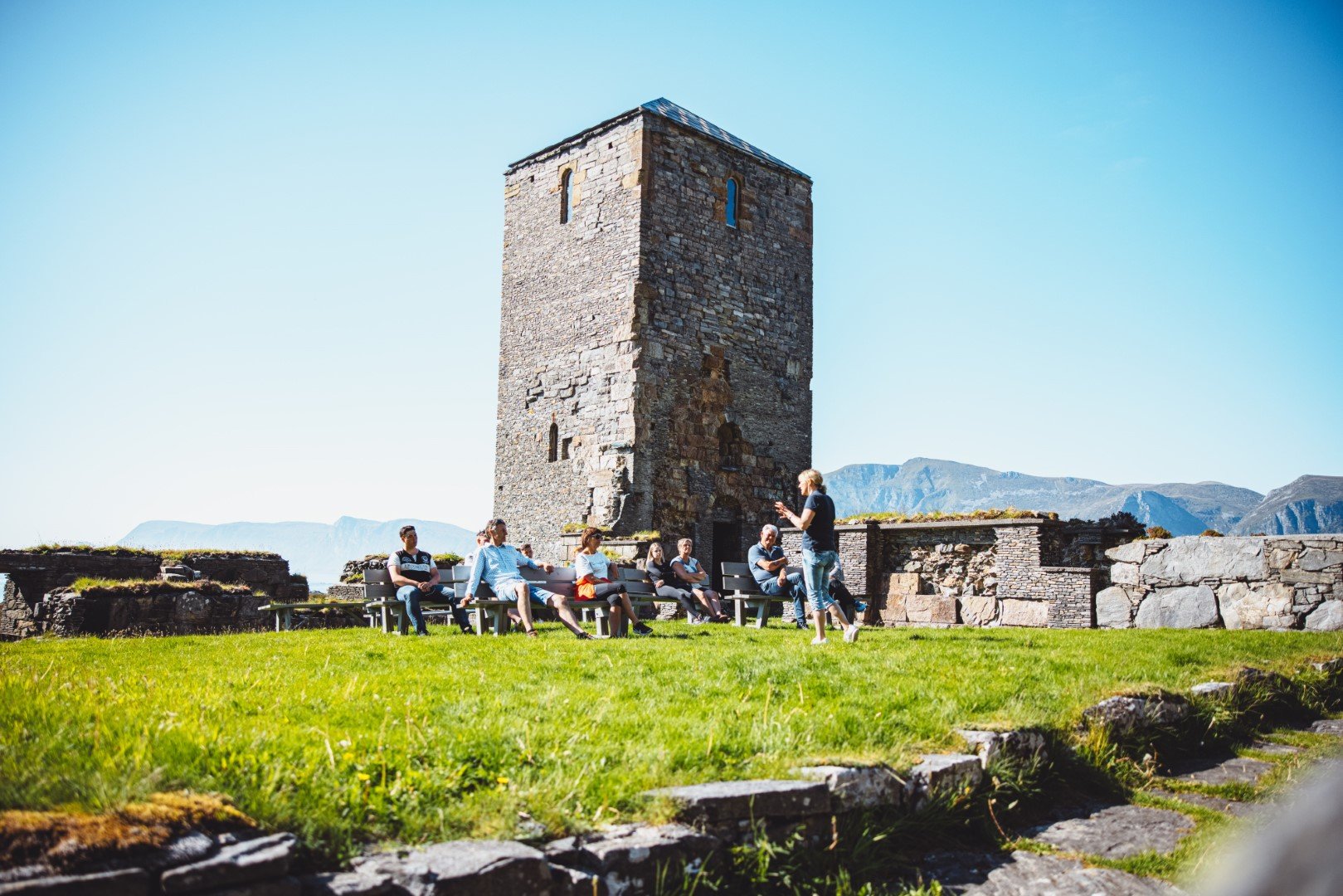Tour guide giving a talk to seated visitors at a historic stone tower on a grassy area, with mountains in the background under a clear blue sky.