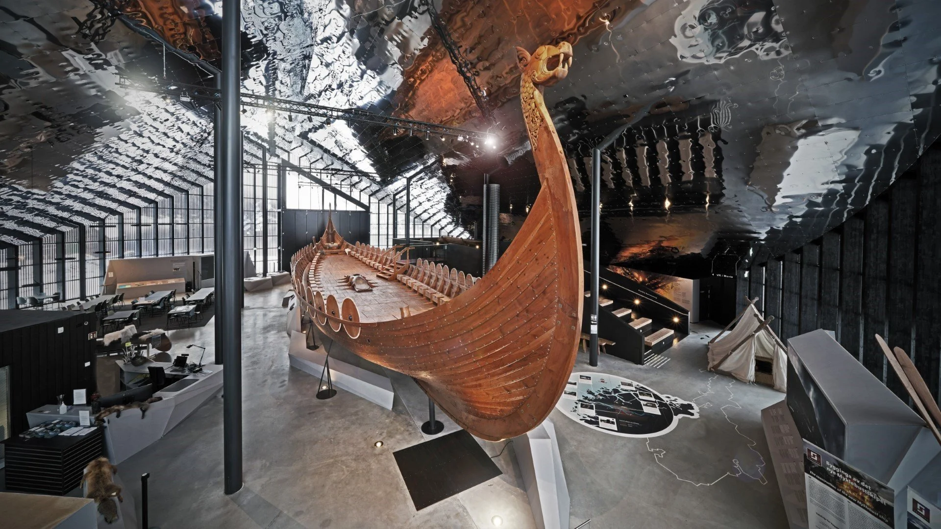 Exhibition hall featuring a large wooden Viking ship replica suspended from the ceiling, with other display elements and informational panels around, in a modern, spacious museum environment.