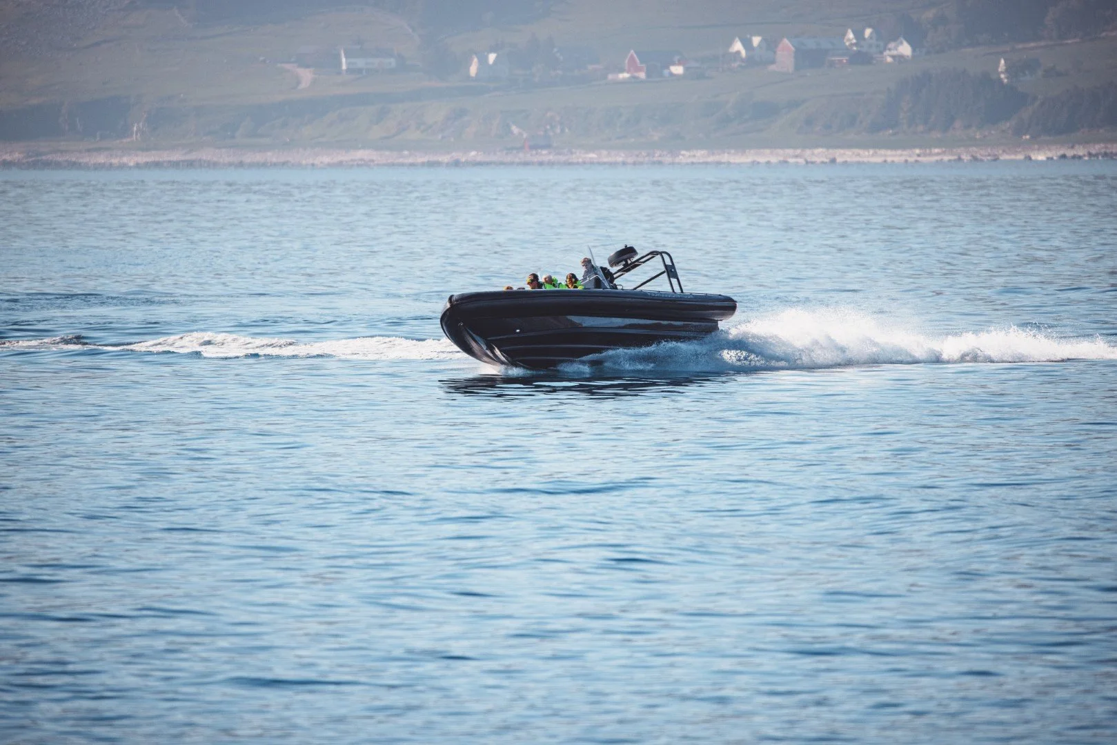 A black speed boat with four passengers cruising on calm water, creating a wake behind it with a scenic coastal background of hills and houses.