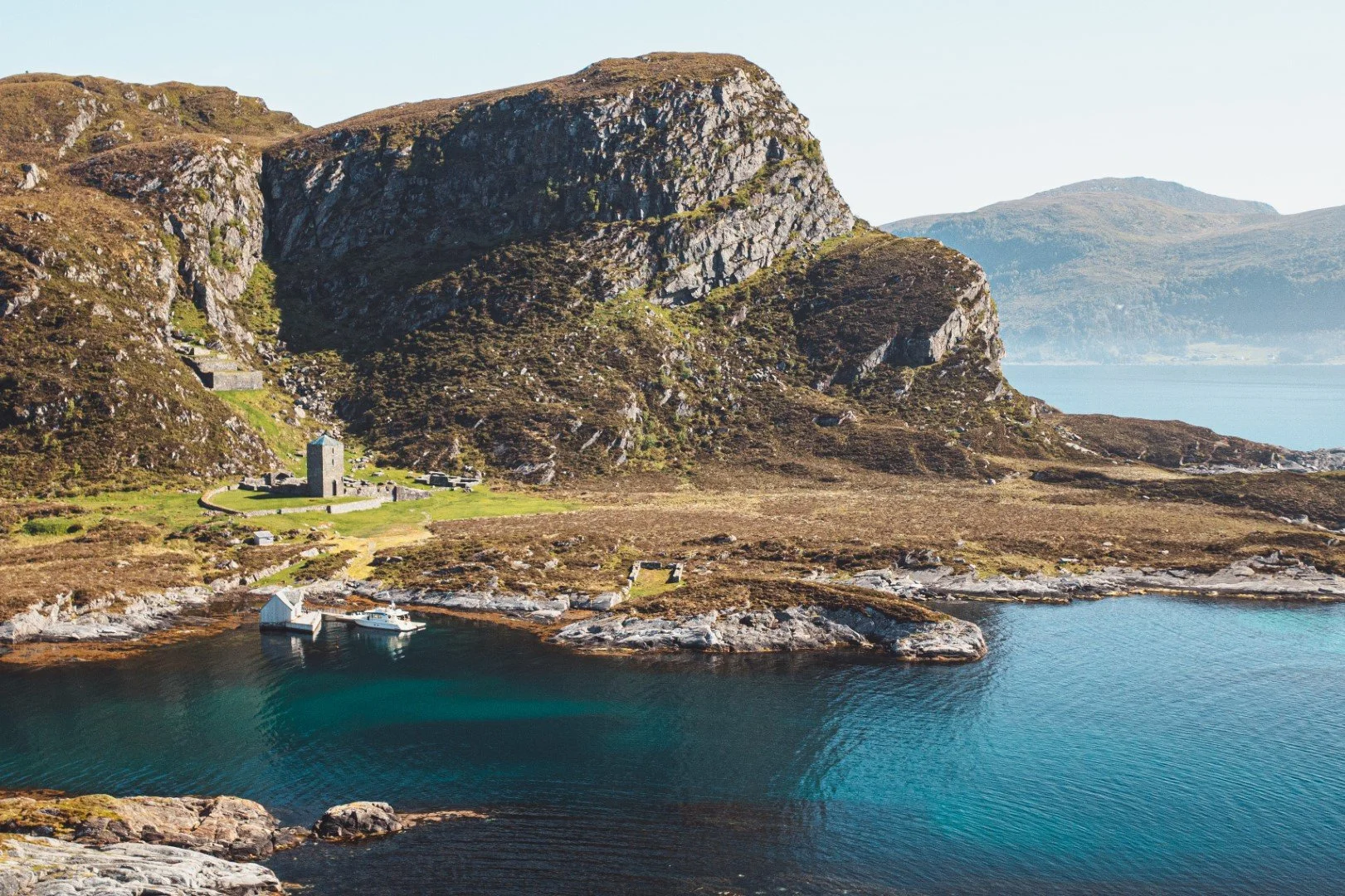 Coastal landscape with rocky shoreline, a small boat house by the water, a white boat docked, green hills, large mountain in the background, and a calm blue body of water.