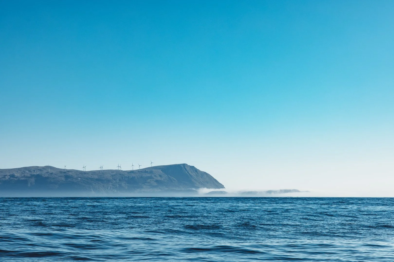 Ocean with calm water and distant landmass with wind turbines on coast under clear blue sky