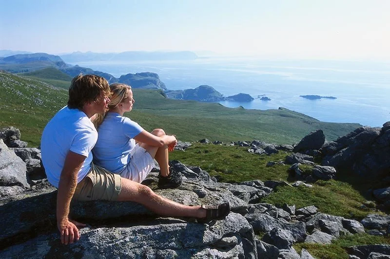 A man and a woman sitting on rocks overlooking a coastal landscape with green hills, rocky formations, and the ocean in the distance.