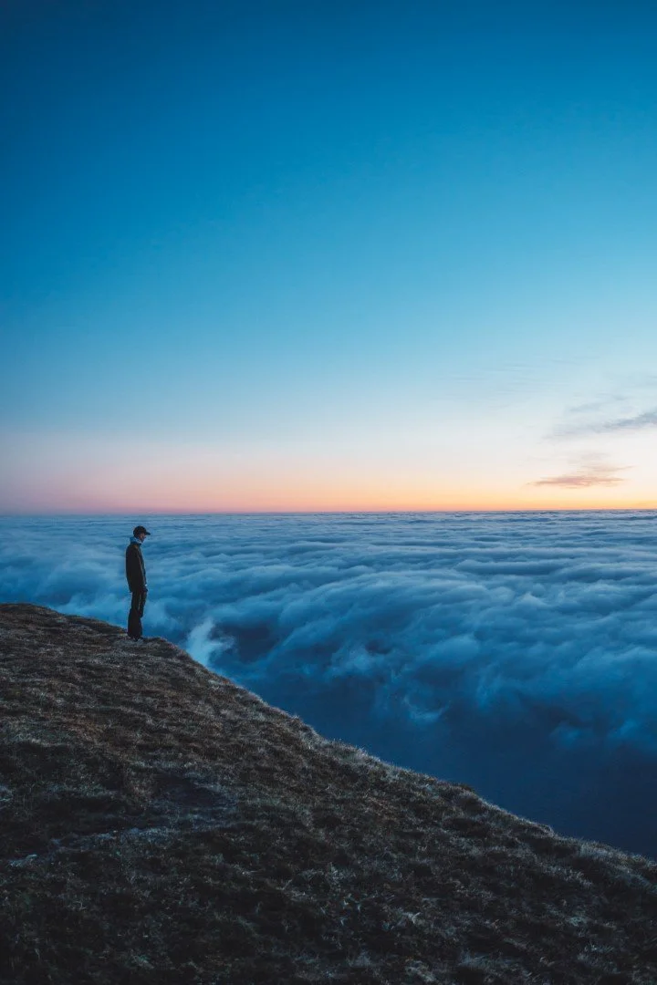 A person standing on the edge of a mountain or hill, looking out over a sea of clouds during sunset or sunrise with a clear sky.