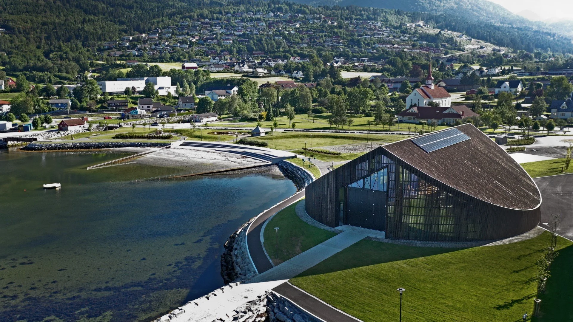 Aerial view of a modern building with a curved dark wooden roof beside a lake, with a park, roads, and a small town in the background.