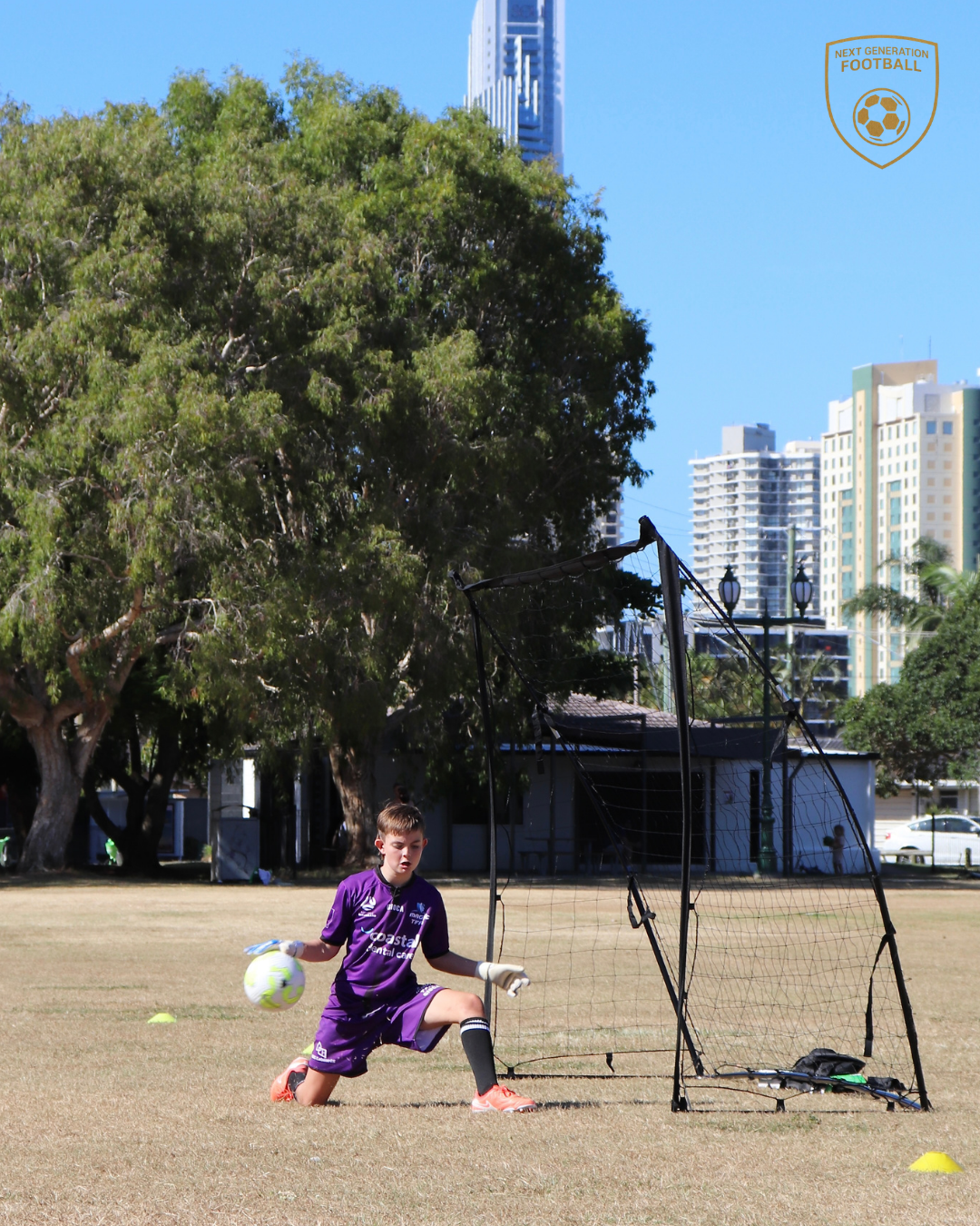 A young soccer player, wearing purple, is practicing goalkeeper skills on a field, kneeling with a soccer ball in hand near a portable goal, with trees, buildings, and a clear blue sky in the background.