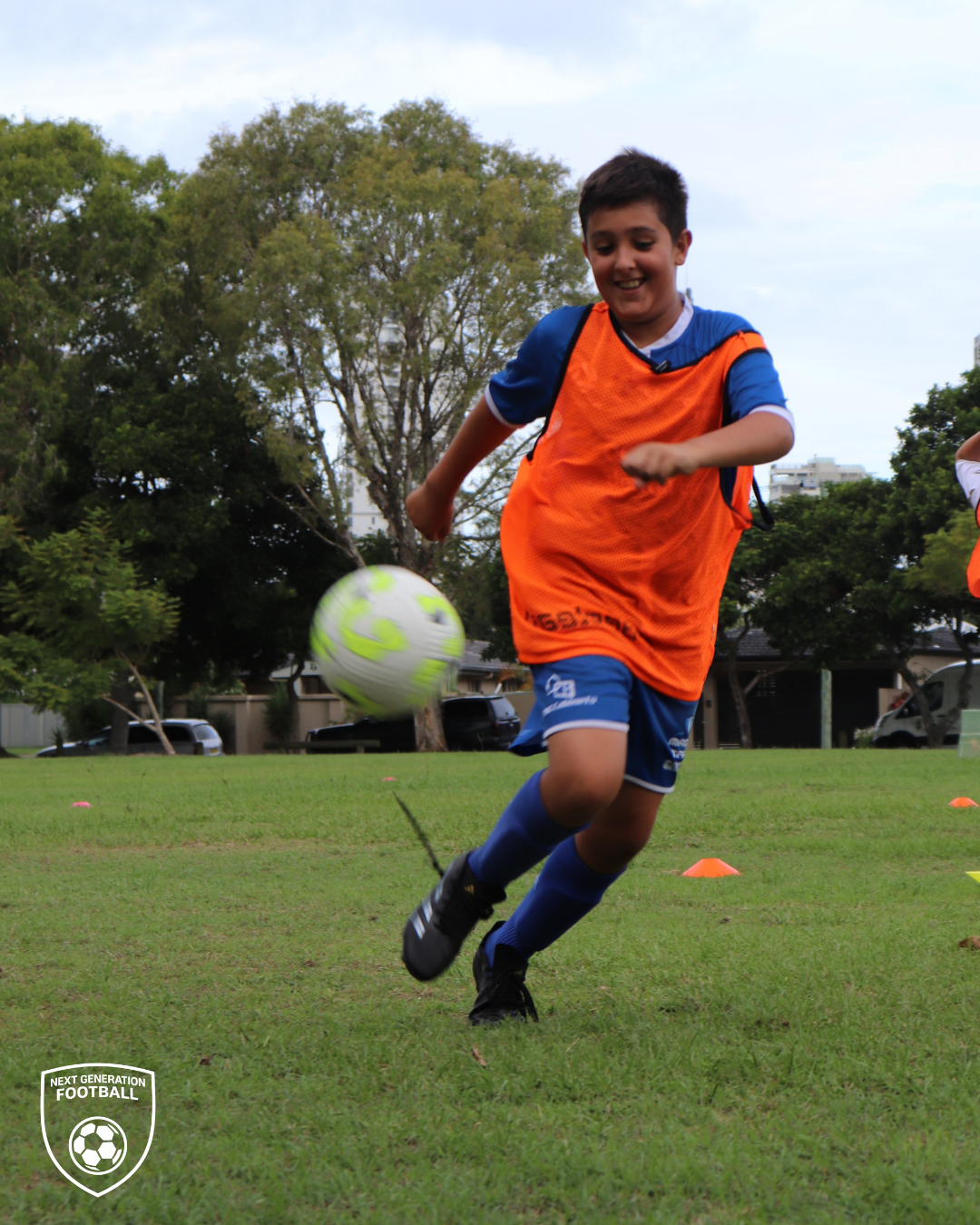 A young boy in a blue sports uniform with an orange training vest is running and kicking a soccer ball on a grassy field during daytime.