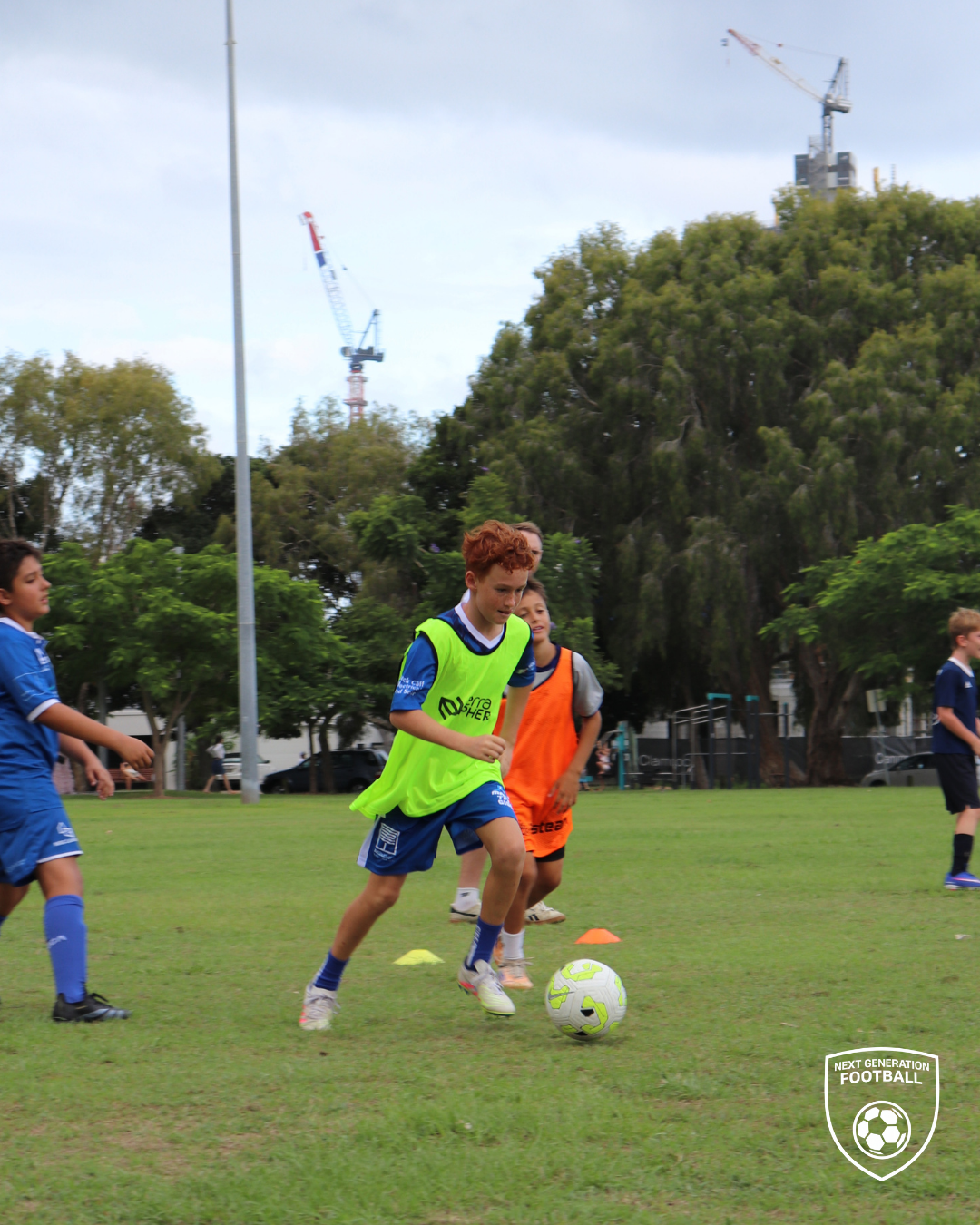 Children playing soccer on a grass field with trees and construction cranes in the background.