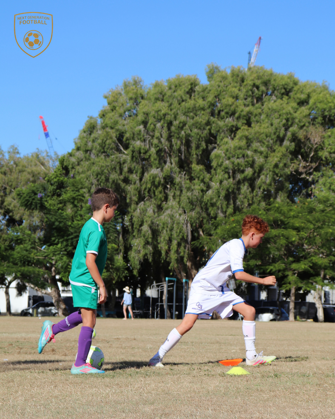 Two young boys practicing soccer on a field with cones, one in a teal uniform and the other in a white uniform, under clear blue sky with trees in the background.