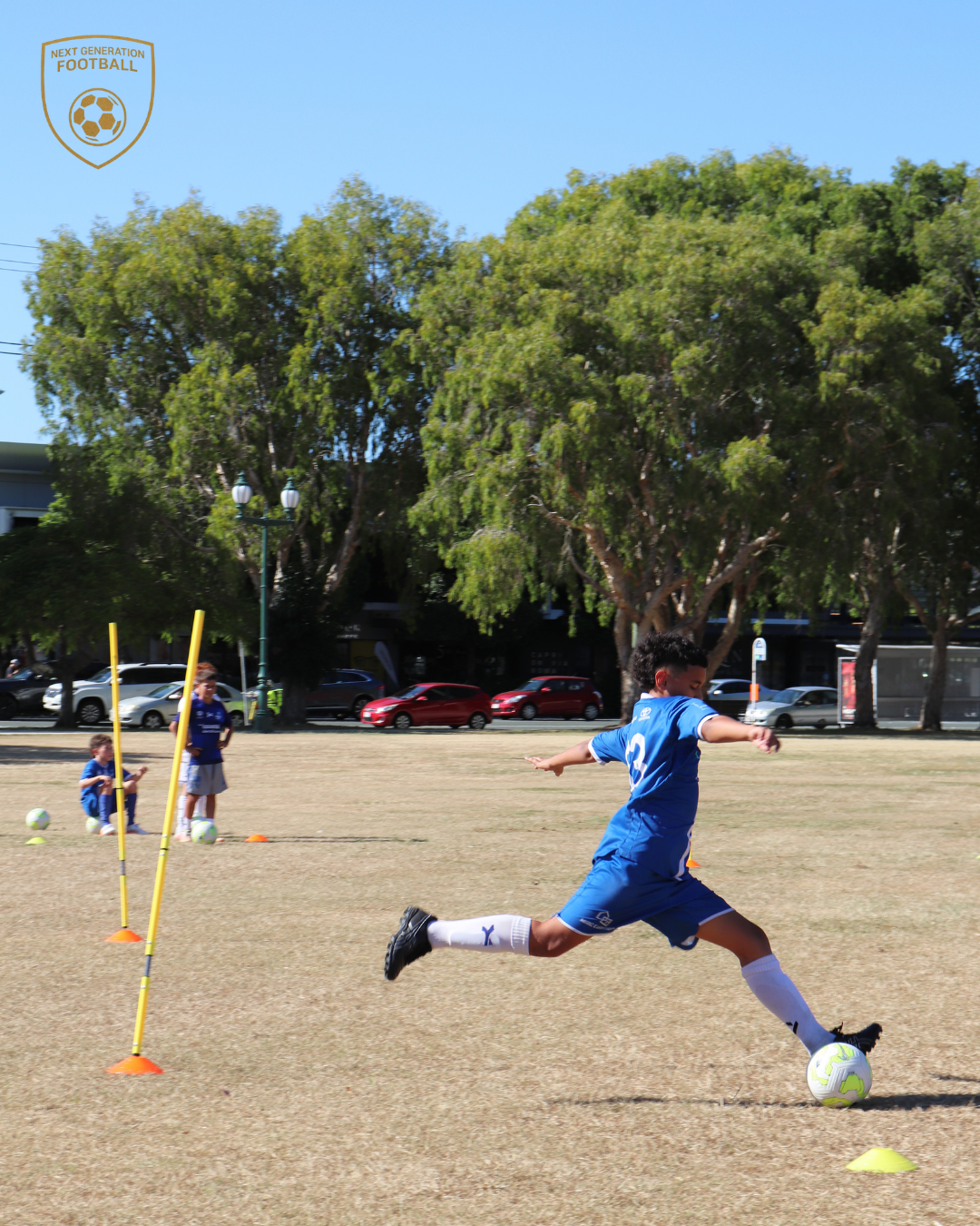 A young boy in a blue soccer uniform kicking a soccer ball on a grassy field during practice, with two other children in similar uniforms near cones and poles, under trees and a parked car background.