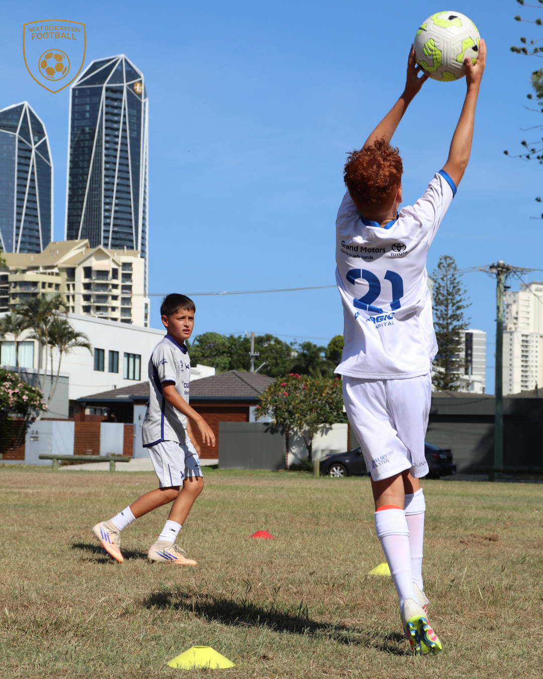 Two young boys in soccer uniforms practicing on a grassy field with city buildings and a clear blue sky in the background.