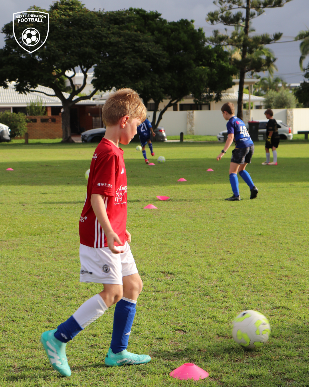 Children participating in a youth soccer practice on a grassy field, practicing dribbling skills around pink cones. One child in red and white kit is closest to the camera, with other kids in blue and black uniforms further in the background. Trees a