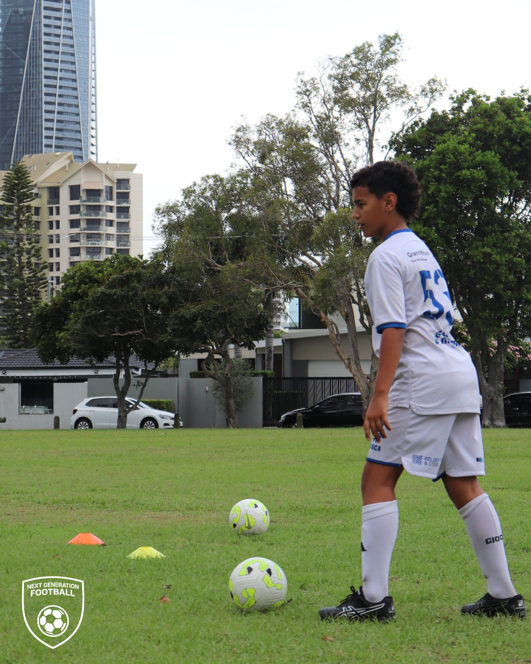 A young soccer player in a white uniform with blue accents practicing on a grass field, standing next to two soccer balls and small orange and yellow cones, with trees, cars, and buildings in the background.