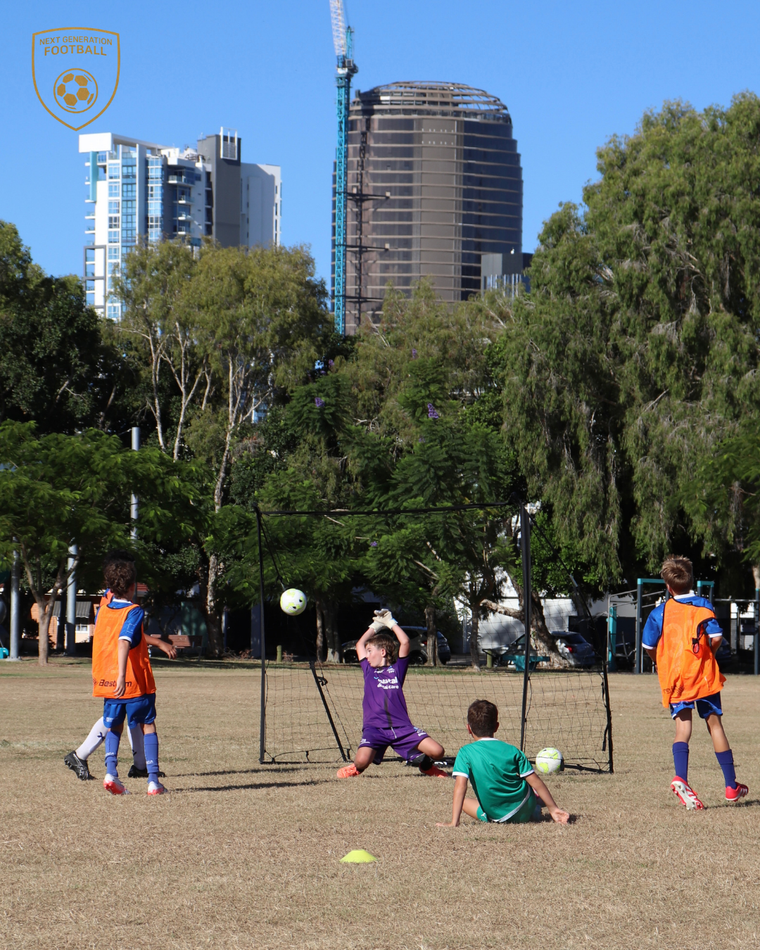 Kids playing soccer on a field with goal net, surrounded by trees and tall buildings in the background.