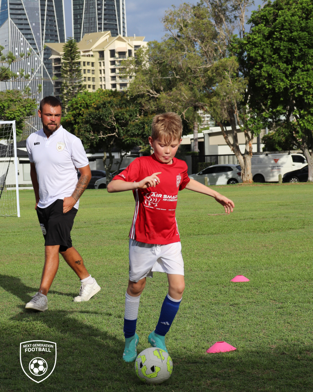 A young boy in a red soccer jersey dribbling a soccer ball on a grassy field during a soccer training session, with an older man observing in the background, and city buildings and trees in the distance.