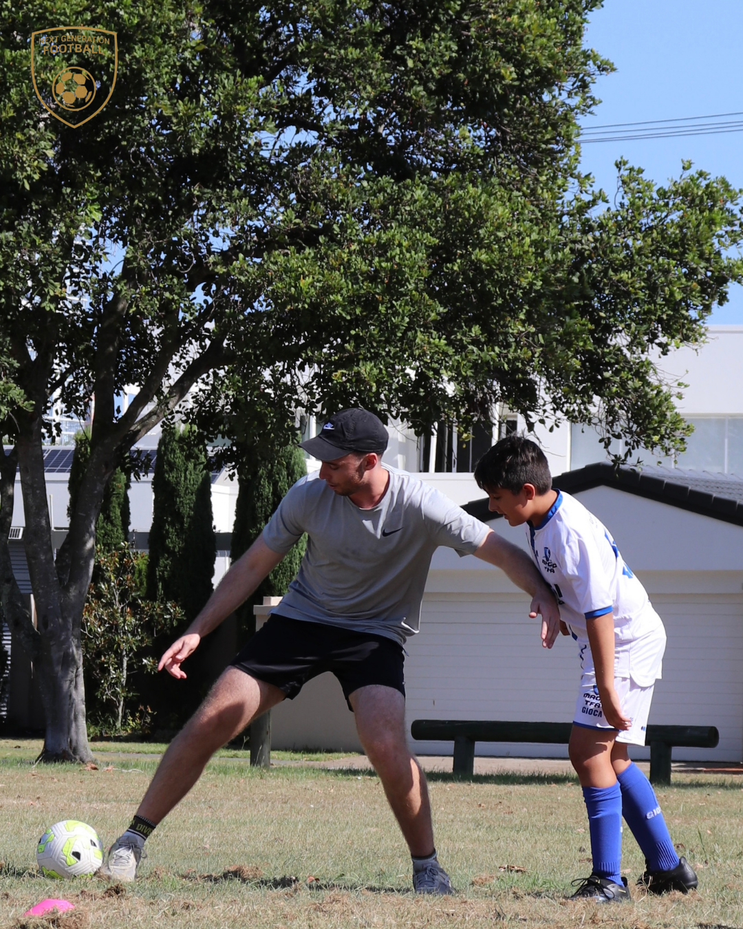 A man and boy practicing soccer outdoors on a grassy field, with trees and a building in the background.