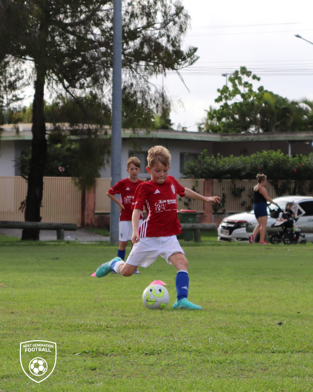 Young boys playing soccer on a grassy field, one about to kick a green and white ball around cones, with spectators including a woman and children in the background.