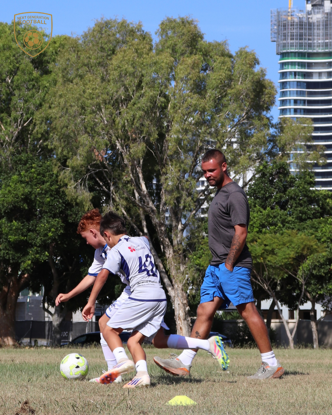 Two young boys playing soccer with a coach or adult man on a field, with trees and tall buildings in the background.