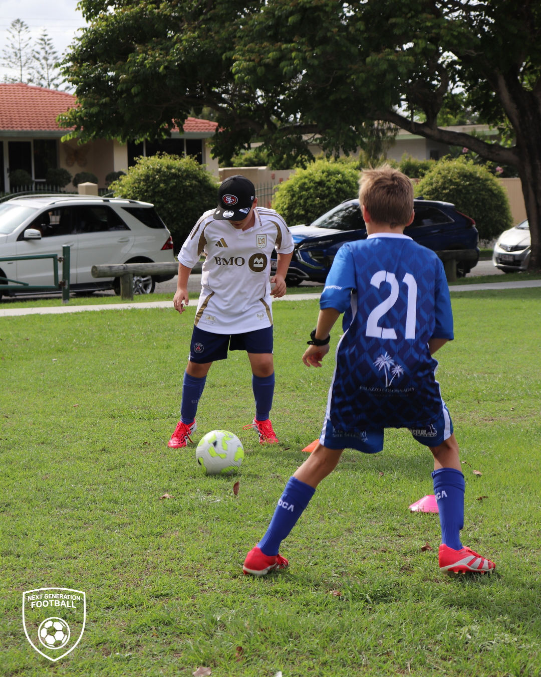Two young boys playing soccer on a grassy field, one wearing a white jersey and black cap, the other wearing a blue jersey with the number 21, surrounded by trees and parked cars.