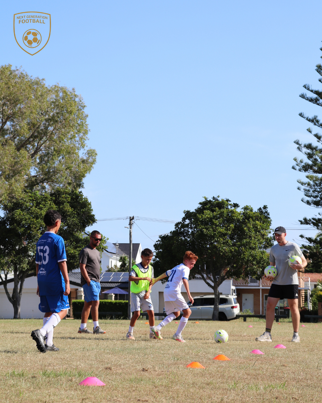 Children participating in a soccer training session outdoors on a sunny day, with a coach holding soccer balls and orange cones placed on the grass.
