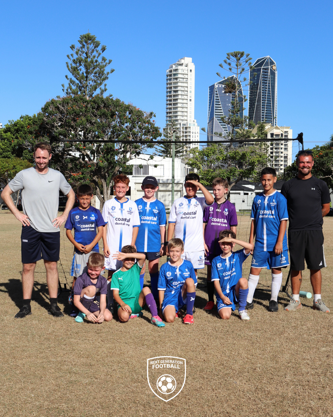 A youth soccer team with two coaches on a soccer field in an urban area with tall buildings, trees, and a clear blue sky in the background.
