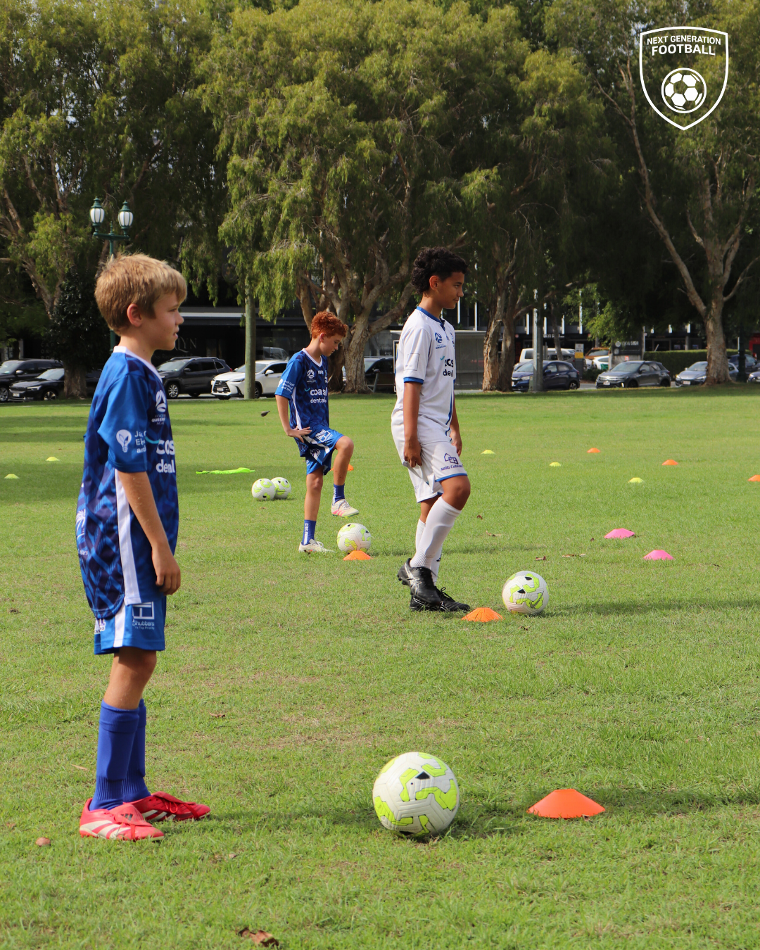 Young boys participating in a soccer training drill on a grassy field, lined up with soccer balls and colorful orange cones, with trees and parked cars in the background.