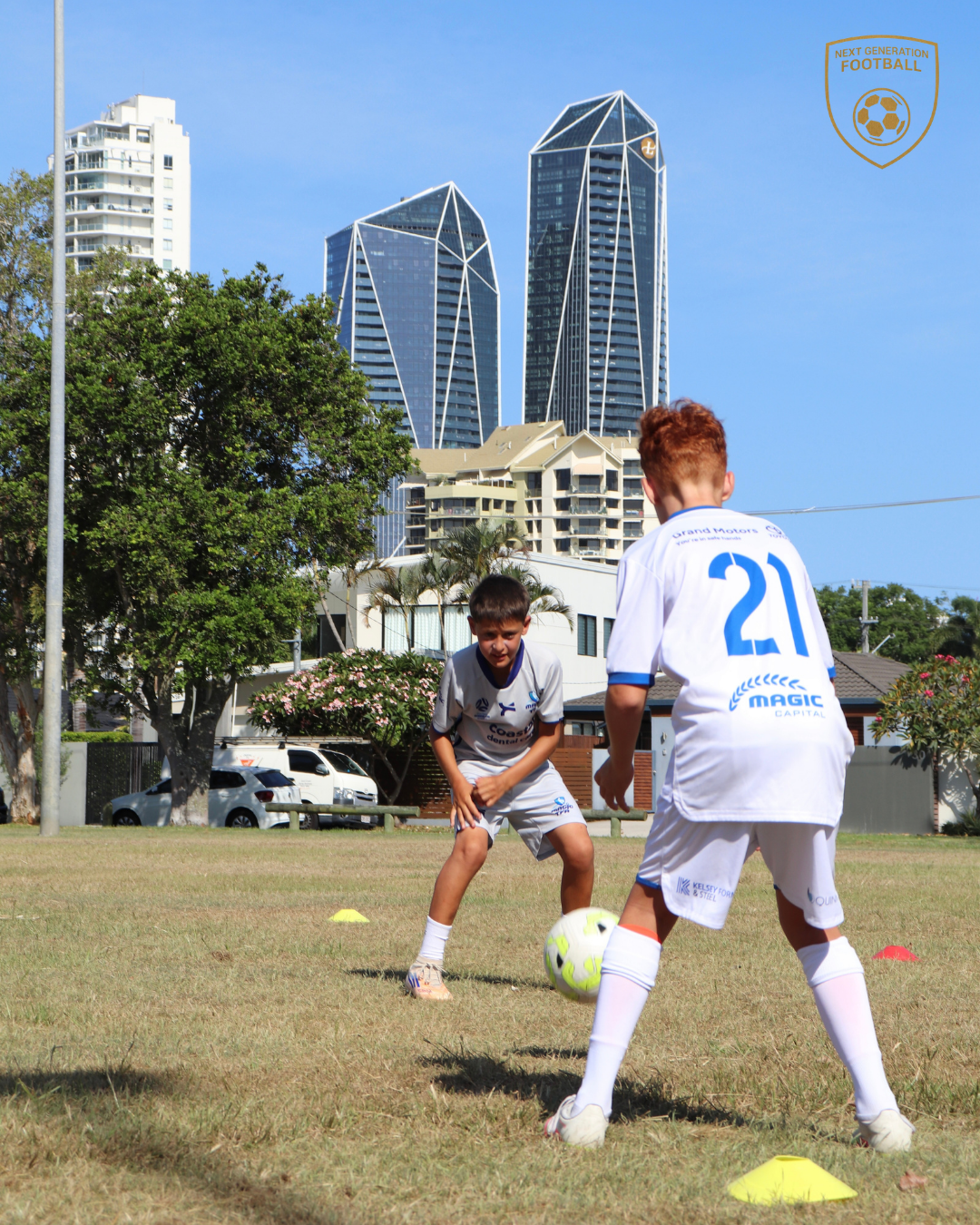 Two young boys playing soccer on a field with yellow cones, city skyscrapers in the background under a blue sky.