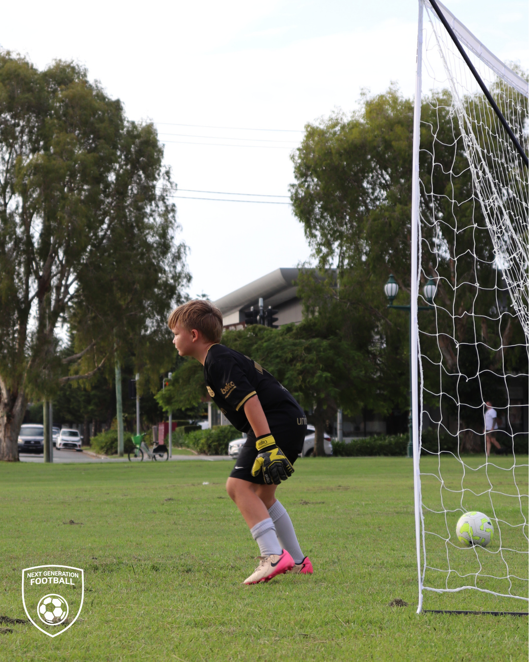A young boy playing soccer as a goalkeeper on a grassy field, standing in front of a goal with a soccer ball inside. Trees and parked cars are visible in the background.
