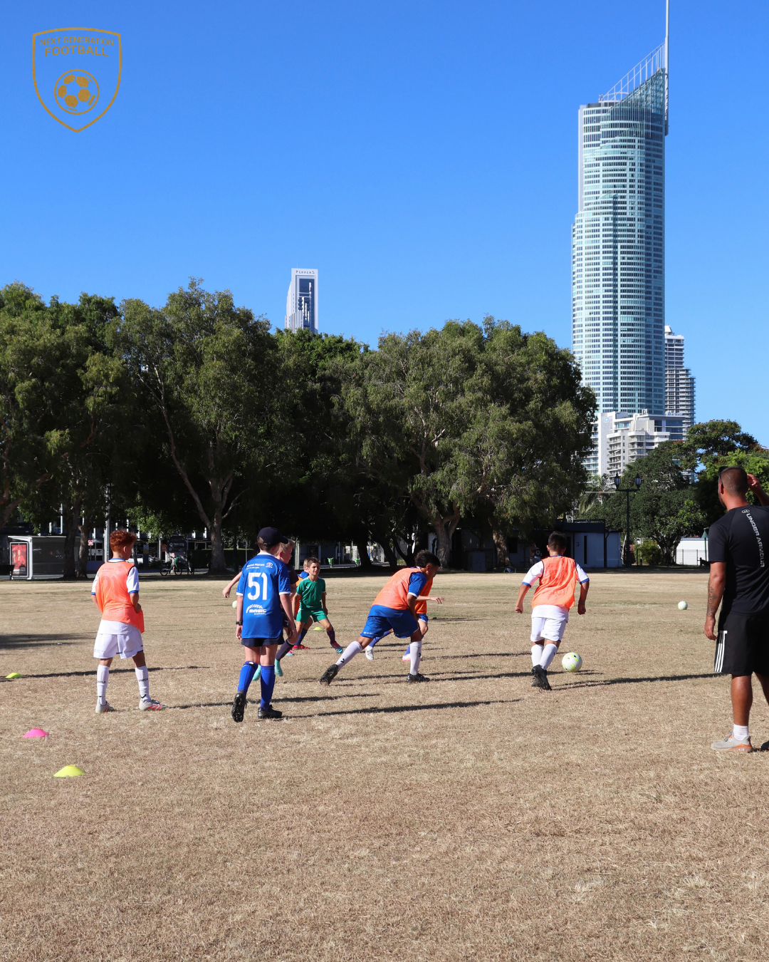 Children playing soccer on a grassy field with tall buildings and trees in the background.