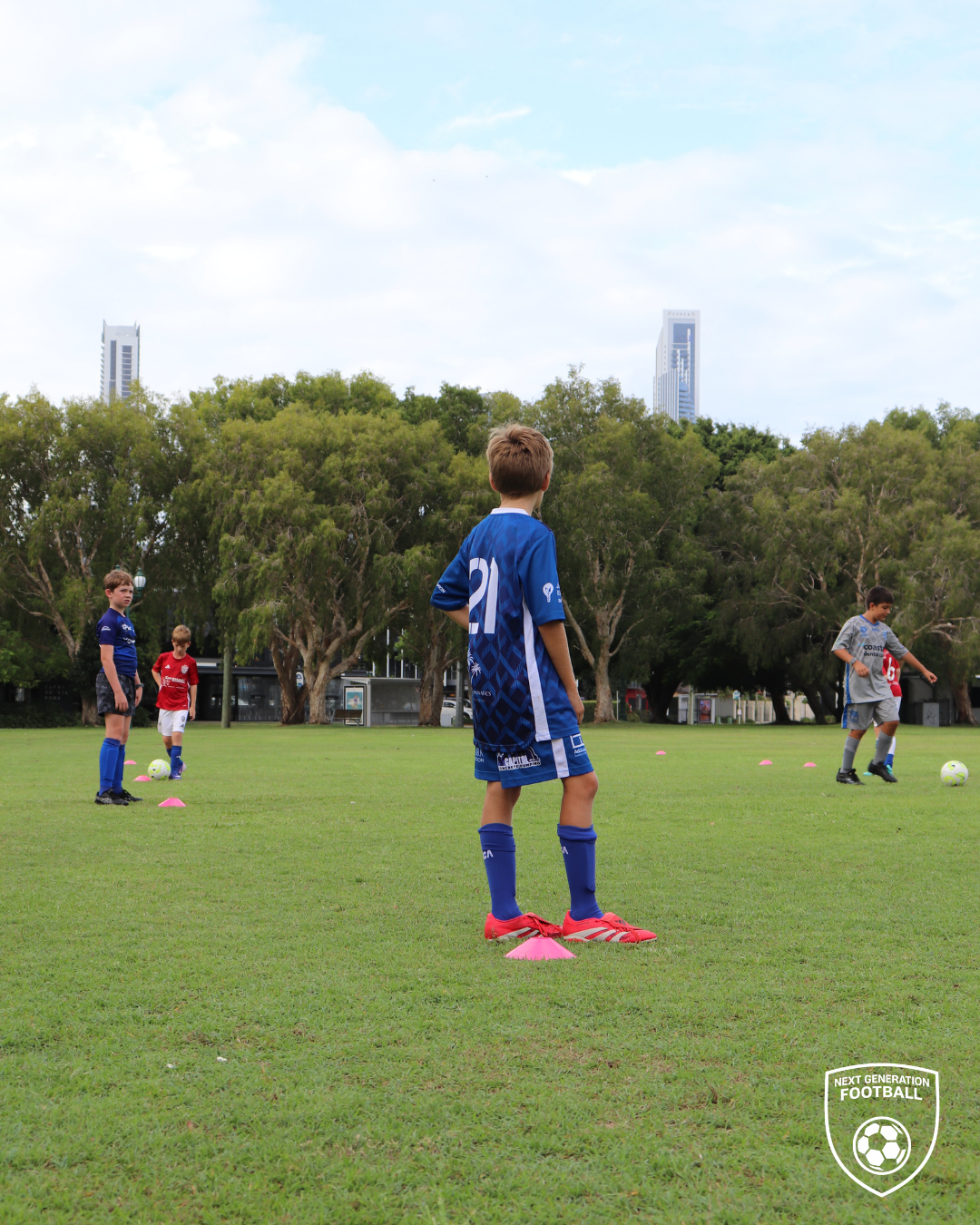 Young boys participating in a youth soccer practice or game on a grassy field, with trees and skyscrapers in the background.