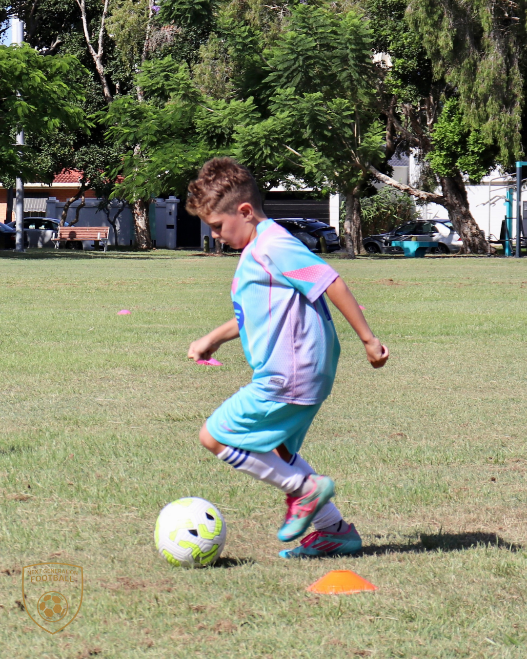 A young boy playing soccer on a grassy field, dressed in sports uniform with a soccer ball and orange cones, surrounded by trees and parked cars in the background.