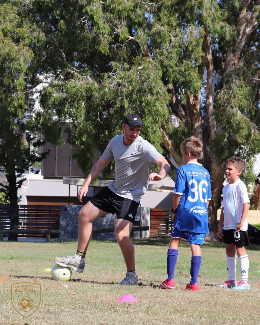 A coach demonstrating soccer skills to two young boys during a practice session outdoors on grass, with cones on the field and trees in the background.