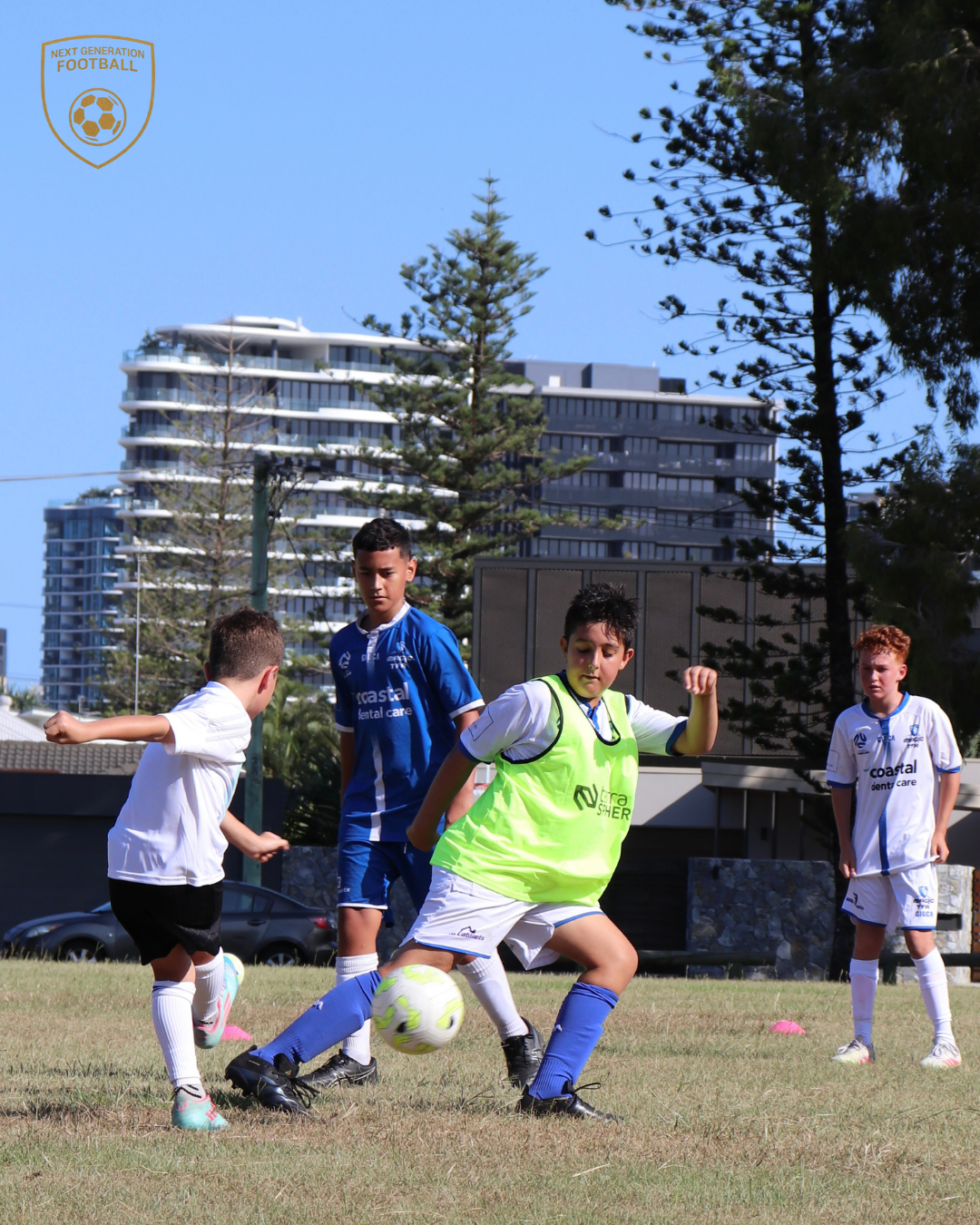 Young boys playing soccer on a field during daytime, with some wearing white jerseys and one in a bright yellow vest. Behind them are trees and modern high-rise buildings.
