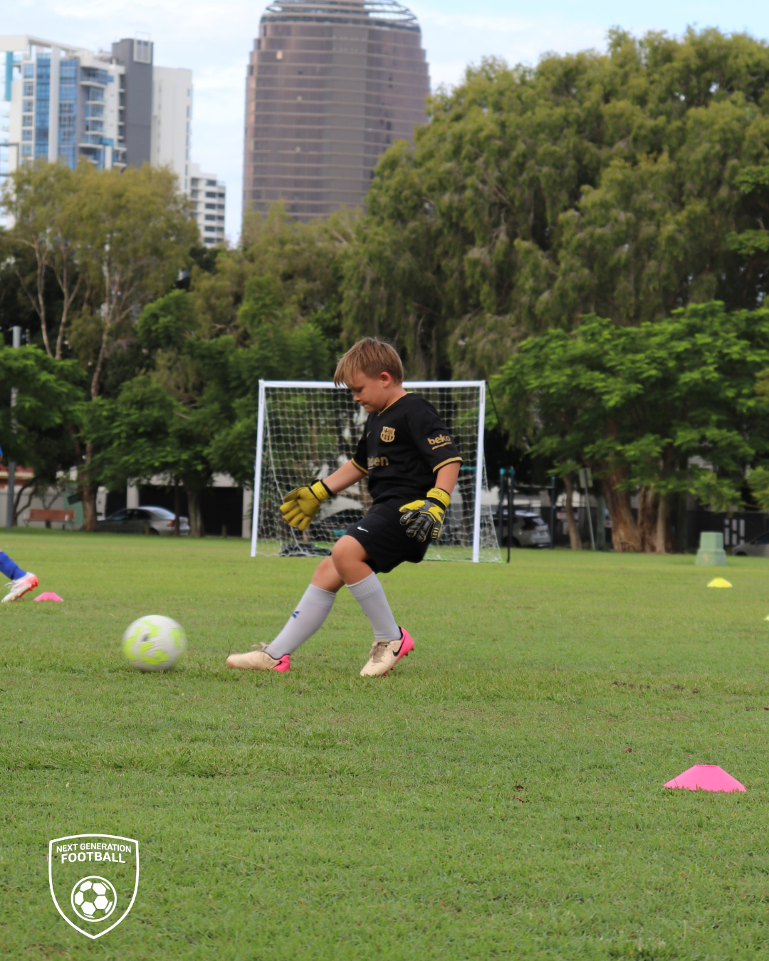 A young boy practicing soccer on a field, wearing a black goalkeeper jersey, shorts, and gloves, kicking a soccer ball near pink cones with a soccer goal in the background, surrounded by trees and city buildings.