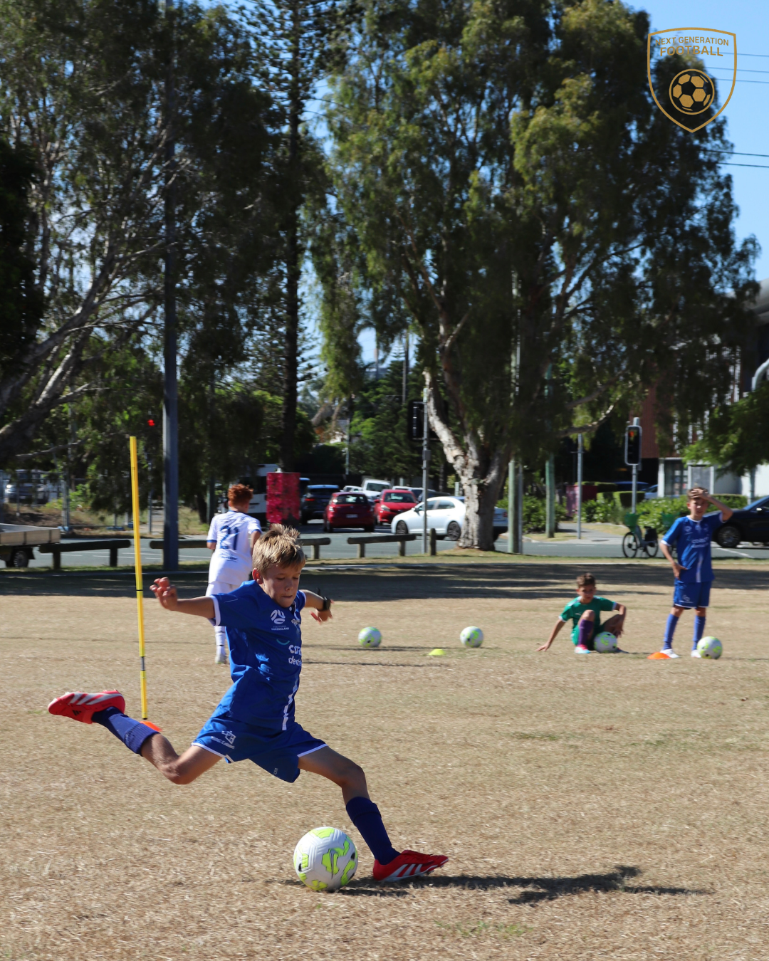 Young boy in blue soccer uniform kicking a soccer ball on a grassy field during practice, with other children and trees in the background.