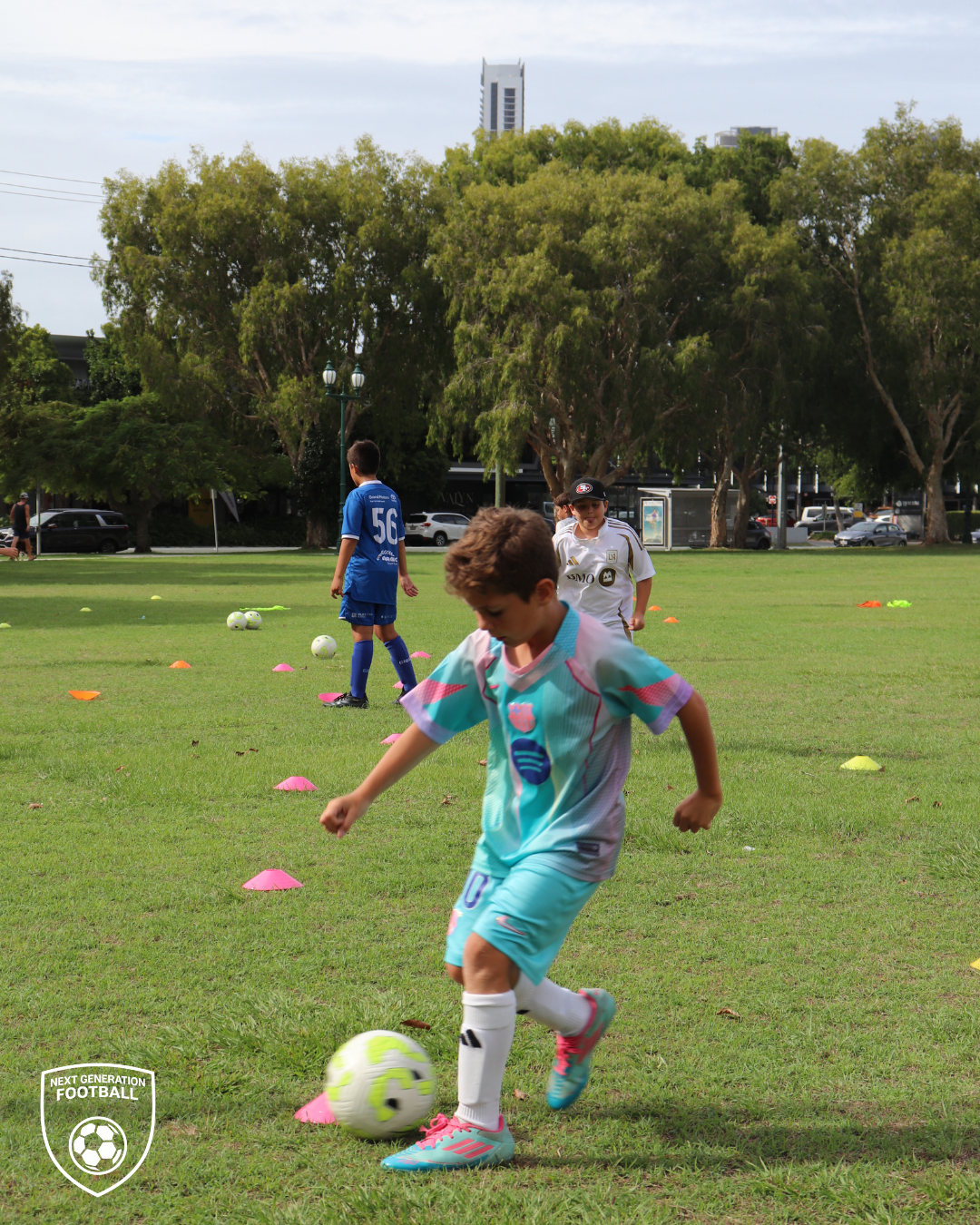 Young boys playing soccer on a grassy field with training cones, with trees and city buildings in the background.